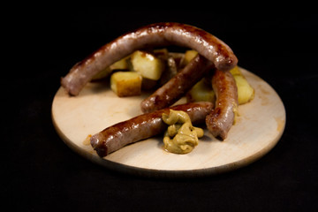 delicious Italian sausage cooked on the grill with browned diced potatoes placed on a wooden cutting board with drops of mustard with a black background 