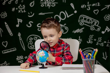 Cute clever boy is sitting at a desk with magnifying glass in hand. Child is reading a book with a blackboard on a background. Ready for school. Back to school