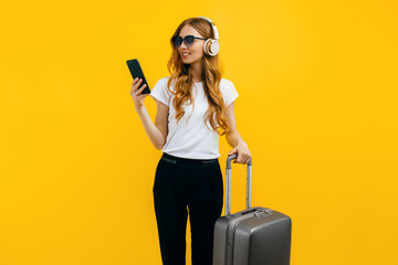 A young smiling tourist, with a suitcase, listening to music on headphones and using a mobile phone, on a colorful yellow background