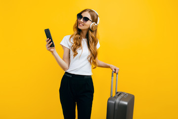 A young smiling tourist, with a suitcase, listening to music on headphones and using a mobile phone, on a colorful yellow background
