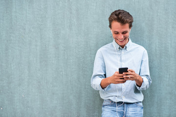 smiling young man leaning against wall with cellphone listening to music with earphones