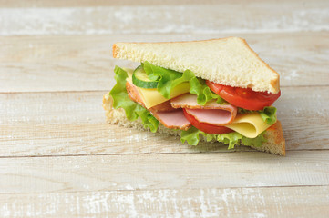 White bread sandwich. The sandwich filling includes cheese, ham, tomato slices, fresh cucumber and salad. Light wooden background. Free space for text.