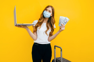 A young woman in a protective medical mask, using a laptop and holding cash dollar bills, on a yellow background. Concept of quarantine, coronavirus