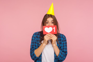 Internet trends. Portrait of positive girl with party cone on head holding social media heart like icon with number one, blogger celebrating first subscriber. studio shot isolated on pink background
