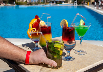 Several multi-colored cocktails on a table by the pool. A glass with a cocktail in hand