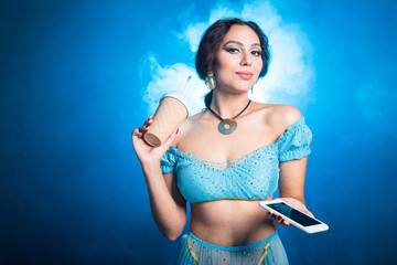 Obraz premium Portrait of young woman in the image of an Eastern fairy Princess in modern clothes holds smartphone and shaker. Carnival and halloween concept.