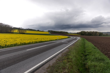 Flowering rapeseed field next to a winding road in the mountains with dramatic clouds in the...