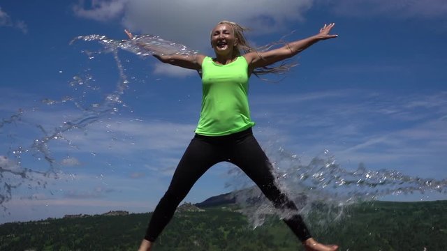 a young woman in a jump with a splash of water. athletic girl jumps up with flying jets of water against the background of summer mountains and sky. Sports and healthy lifestyle. Pretty fit girl