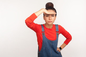 Portrait of girl in denim overalls holding hand over eyes and looking at long distance with confident determined expression, being ambitious about far future. studio shot isolated on white background