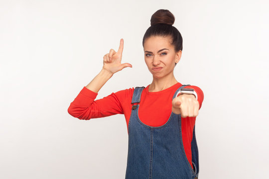 Portrait of irritated girl with hair bun in denim overalls showing loser gesture and pointing to camera, blaming for defeat, naming you unlucky jobless. indoor studio shot isolated on white background