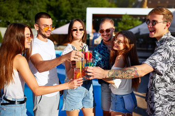 Group of friends having fun at poolside summer party clinking glasses with summer cocktails on sunny day near swimming pool. People toast drinking fresh juice at luxury villa on tropical vacation. - obrazy, fototapety, plakaty