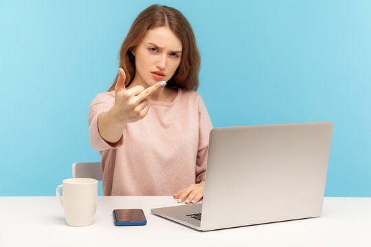 Impolite Furious Young Woman Employee Sitting At Workplace With Laptop, Showing Middle Finger, Expressing Disrespect And Hate, Conflict With Coworkers. Indoor Studio Shot Isolated On Blue Background