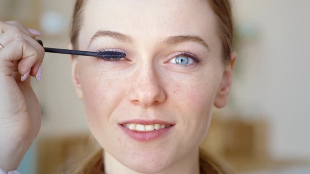 Confident happy young woman holding mascara apply on eyeleashes prepare getting ready in the morning, smiling beautiful girl doing make up put cosmetics on face looking in mirror in bathroom at home