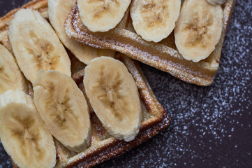 Belgian waffles with banana and sugar powder on dark background.