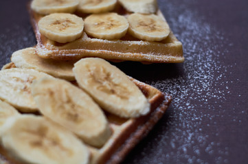 Belgian waffles with banana and sugar powder on dark background.
