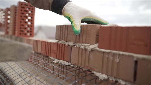 bricklayer laying, well, bricks to make a wall, he is pulling grout out of a joint with his trowel. This man is really working hard. Worker builds a house of brick.