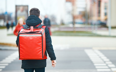 Delivery boy walks with red thermal bag on city street. Fast and convenient food delivery service for self-isolated citizens during an epidemic of viral disease. Boy delivering food to customers