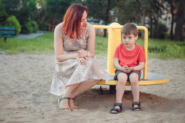 Mom and son on playground. Boy in bright red T-shirt and young woman in dress outdoors summer. Everyday life. Concept of childhood and relationship of children with parents