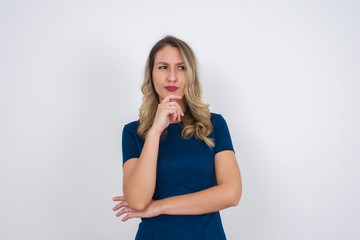 Shot of contemplative thoughtful Young woman keeps hand under chin, looks thoughtfully upwards, dressed in casual clothes, poses over white background with free space for your text