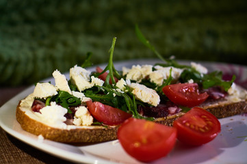 Large toast of rye bread with pieces of cheese, arugula, cherry tomatoes and sausage.