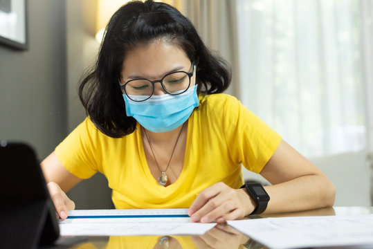 Young Woman Architect designer wear eyeglasses and yellow shirt and blue surgical mask working on blueprints for interior design project during quarantine in pandemic Coronavirus or Covid 19