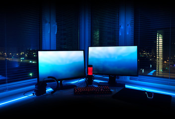 Dark room for a gamer with a computer table, neon lights. Gamezone. Stream room with devices, reflection in the window of the night city, lights. Modern interior for a teenager. © MiaStendal