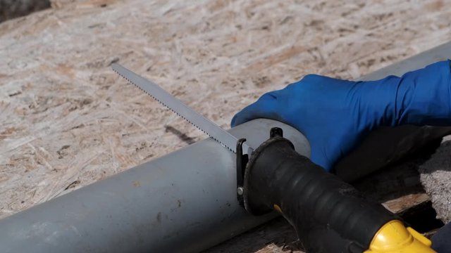 Plumber man in rubber gloves, cuts the plastic sewer pipe, electric saber saw. Close-up.