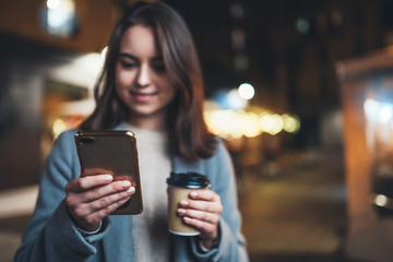 hipster girl drink coffee outdoors using mobile smartphone on background bokeh light in night city, woman hold in hands sellphone, online wi-fi internet