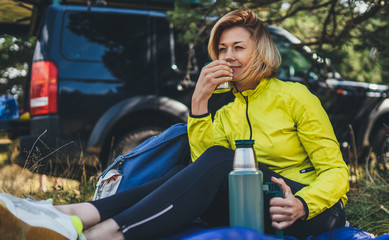Girl smiles and enjoys summer nature hold hand mug of warm tea during recreation trip, hiker tranquil drink coffee from thermos in green forest tourist traveling auto camping. woman emotion outdoor
