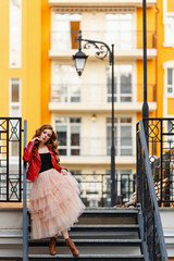 Sad woman weared in tulle skirt and red leather jacket in the City near old classic lantern. Fashion style girl with long curled red hair. Street Modern urban style. Young curled girl in a big City