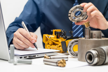 Professional mechanic engineer inspecting valve plate of hydraulic piston pump and writing reports for during working day in office, repair maintenance heavy machinery concept