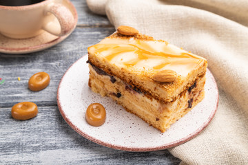 Honey cake with milk cream, caramel, almonds and a cup of coffee on a gray wooden background. Side view.