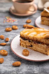 Honey cake with milk cream, caramel, almonds and a cup of coffee on a gray wooden background. Side view, selective focus.