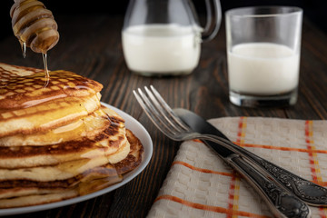 Honey is poured onto a high stack of golden pancakes laid on a gray plate, flows down the edges, on...