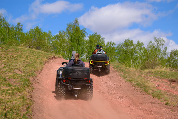 Young people with girls ride ATVs. Mountainous terrain. Dusty road is red. View from the back. Concept of adventure, tourism.