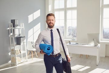 Happy successful businessman in a white shirt with yoga mat standing in the office.