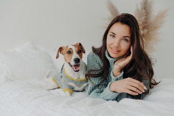 Horizontal shot of pleasant looking tender woman spends free time with favorite pet, looks directly at camera, enjoys domestic atmosphere, cares about dog. People, animals and friendship concept