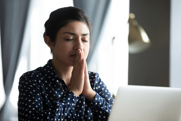 Head shot hopeful young indian student praying god before starting passing online test examination on computer. Focused millennial hindu woman asking for help good luck, sitting at table with laptop. - obrazy, fototapety, plakaty