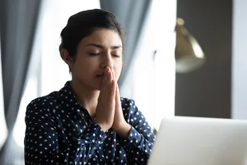 Tuinposter Head shot hopeful young indian student praying god before starting passing online test examination on computer. Focused millennial hindu woman asking for help good luck, sitting at table with laptop. © fizkes