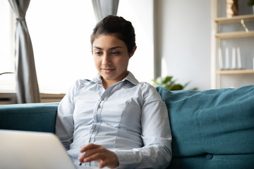 Calm young indian woman freelancer sitting on sofa with computer, working remotely from home. Pleasant millennial girl student study online, watching educational lecture on laptop, sitting on sofa.