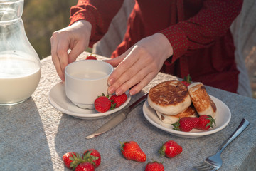 Female hands hold a mug with milk on a table with cheesecakes .