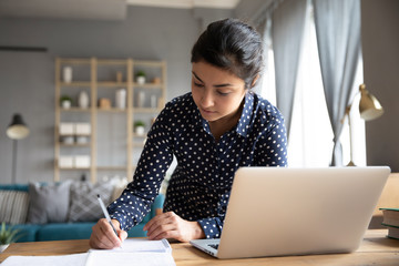 Focused young indian student writing notes from online seminar lecture workshop, standing at table, looking at computer screen. Concentrated millennial hindu ethnic woman doing research at home.