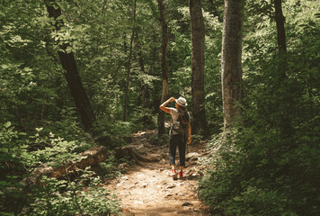 woman walks in the summer forest with a backpack and a hat. Stands and looks at the opening of sunlight.