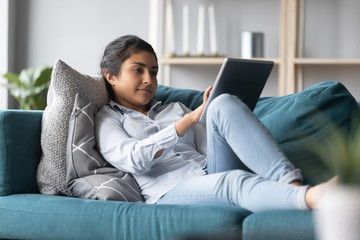 Full length focused pleasant young indian woman lying on comfortable sofa, using digital computer tablet. Happy millennial hindu girl resting on couch, chatting with friends, chatting alone at home.