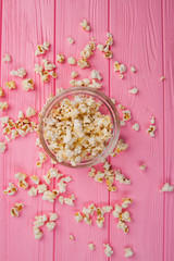 Flat lay popcorn in a glass bowl and sprinkled popcorn around it. Pink wooden desk.