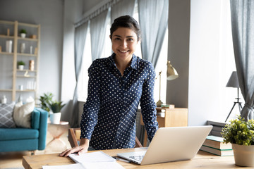Smiling young indian woman freelancer standing at table with computer, looking at camera. Pleasant attractive hindu female student posing for photo in modern living room, ready for study at home.
