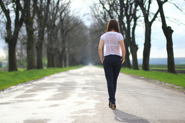 Back view of a young girl walking on the road. Tall trees along the way