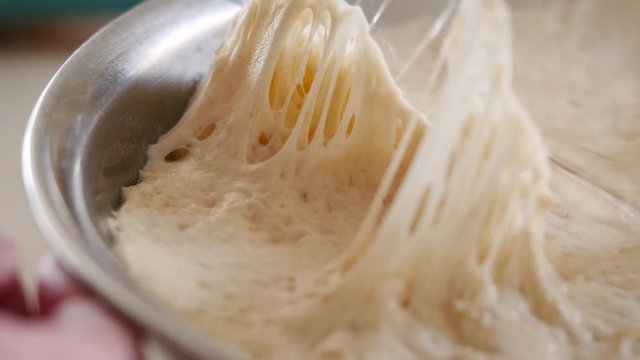 Pulling plastic wrap off risen bread dough in a metal mixing bowl. The gluten stretches and the raw flour mixture can be formed into a loaf and baked. Making sourdough is the hot trend of lockdown.
