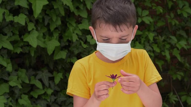 a small child in a medical mask tears off the petals of a flower, a boy in a yellow t-shirt guesses on a flower, tearing the petals from it, against the background of ivy