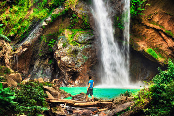Happy man backpacker enjoying amazing tropical waterfall raised hands Travel Lifestyle and success...